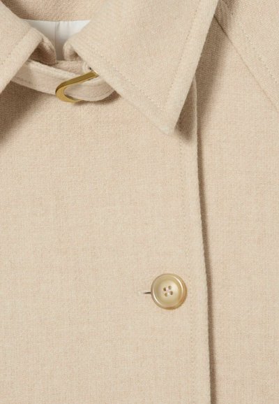 Cappotto di lana beige con ampio colletto, una fascia con bottoni fissata da una fibbia color oro sullo scollo e un unico bottone visibile.