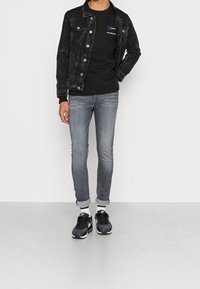 Černá džínová bunda přes černé tričko, šedé slim-fit džíny, bílé ponožky a černé tenisky s bílými akcenty. Neutrální pozadí.