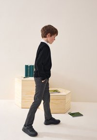 Garçon en pull noir et pantalon gris marche sur un sol clair. Baskets noires. Bloc hexagonal en bois et carnets verts en arrière-plan.