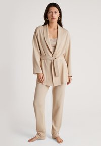 Beige loungewear set met een wikkelstijl jas met een strikceintuur en brede broekspijpen, gemaakt van zachte, textuur stof. Opvallende kantdetails aan de binnenkant van de top.