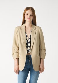 Beige Blazer mit tailliertem Design, ausgestattet mit zwei vorderen Taschen, Reverskragen und hochgerollten Ärmeln, getragen über einem Oberteil mit Blumenmuster.