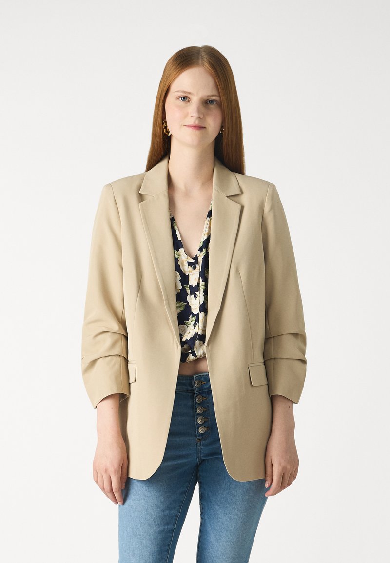 Beige Blazer mit tailliertem Design, ausgestattet mit zwei vorderen Taschen, Reverskragen und hochgerollten Ärmeln, getragen über einem Oberteil mit Blumenmuster.