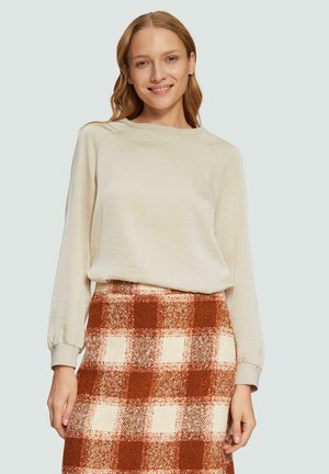 Haut beige à manches longues avec des manches raglan, associé à une jupe à carreaux présentant des carrés marron et crème dans un tissu texturé.