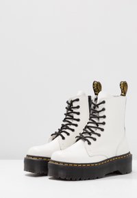 Botas de tobillo de cuero blanco con cordones negros, suela negra gruesa, costuras amarillas y lengüetas. Superficie texturizada y diseño robusto.