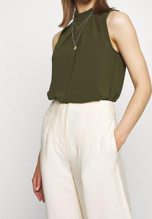 Woman wearing sleeveless olive green top with high neck and layered necklaces, paired with high-waisted cream wide-leg pants, hand in pocket.