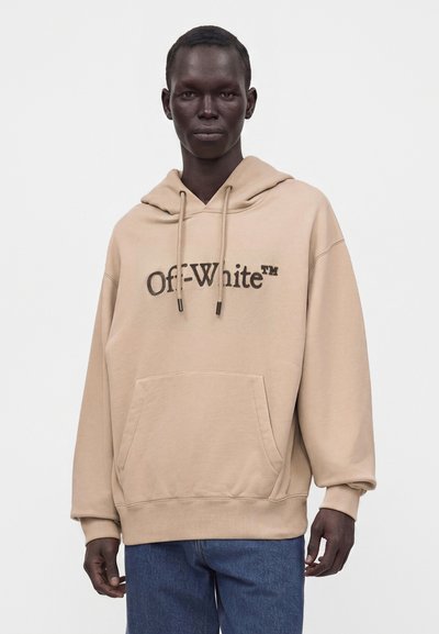 Personne noire portant un sweat à capuche beige Off-White avec poche avant et jean bleu, debout devant un fond blanc uni.
