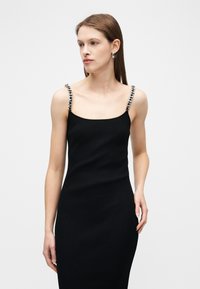 Γυναίκα με μαύρο αμάνικο φόρεμα με λουριά αλυσίδας, που στέκεται μπροστά σε απλό φωτεινό φόντο, κοιτάζοντας ελαφρώς προς τα αριστερά.