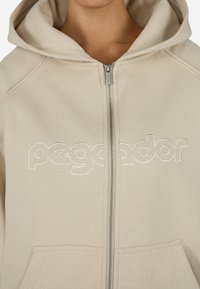 Beige színű cipzáras kapucnis pulóver, húzózsinórral, fehér hímzett "pegador" felirattal a mellrészen és elülső zsebekkel.