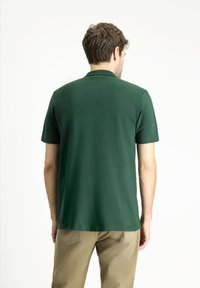 Polo a maniche corte in tessuto verde scuro, con colletto classico e una trama liscia, indossato con pantaloni beige chiaro.