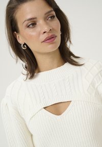 White knitted sweater featuring textured patterns, a high neckline, and a v-cut opening at the chest. Model wears silver hoop earrings.