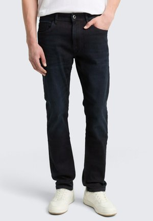 Man in slim-fit donkere jeans en witte sneakers, staand met één hand in zijn zak tegen een effen achtergrond.