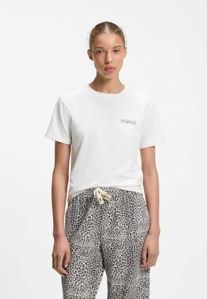 Ung kvinde med trukket hår bærer en hvid T-shirt med lille logo og leopardprint bukser med mærket snøre.