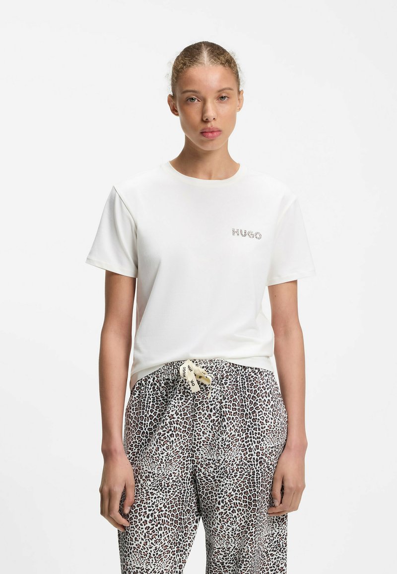 Junge Frau mit zurückgebundenem Haar trägt ein weißes T-Shirt mit kleinem Logo und Leoparden-Print-Hose mit markiertem Kordelzug.