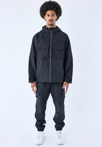 Conjunto de chaqueta y pantalones negros, hecho de una tela suave. La chaqueta presenta un cierre con cremallera y dos bolsillos en el pecho. Los pantalones tienen bolsillos tipo cargo y puños ajustados.