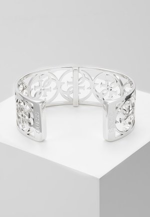 Zilveren manchetarmband met opengewerkte bloemenpatronen en de merknaam "GUESS" gegraveerd aan beide uiteinden, getoond op een wit voetstuk.