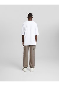 Valkoinen oversize-t-paita, lyhyet hihat, yhdistetty beigeihin cargo-housuihin, joissa sivutaskut, päällä valkoiset tennarit neutraalilla taustalla.