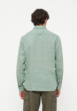 Uomo con camicia verde chiaro a maniche lunghe e pantaloni cargo verde oliva, visto da dietro su uno sfondo bianco uniforme.