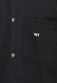Camisa preta de botões feita de um tecido texturizado, com um bolso no lado esquerdo do peito onde está bordado "OBEY" em branco. Botões de latão acrescentam detalhe.