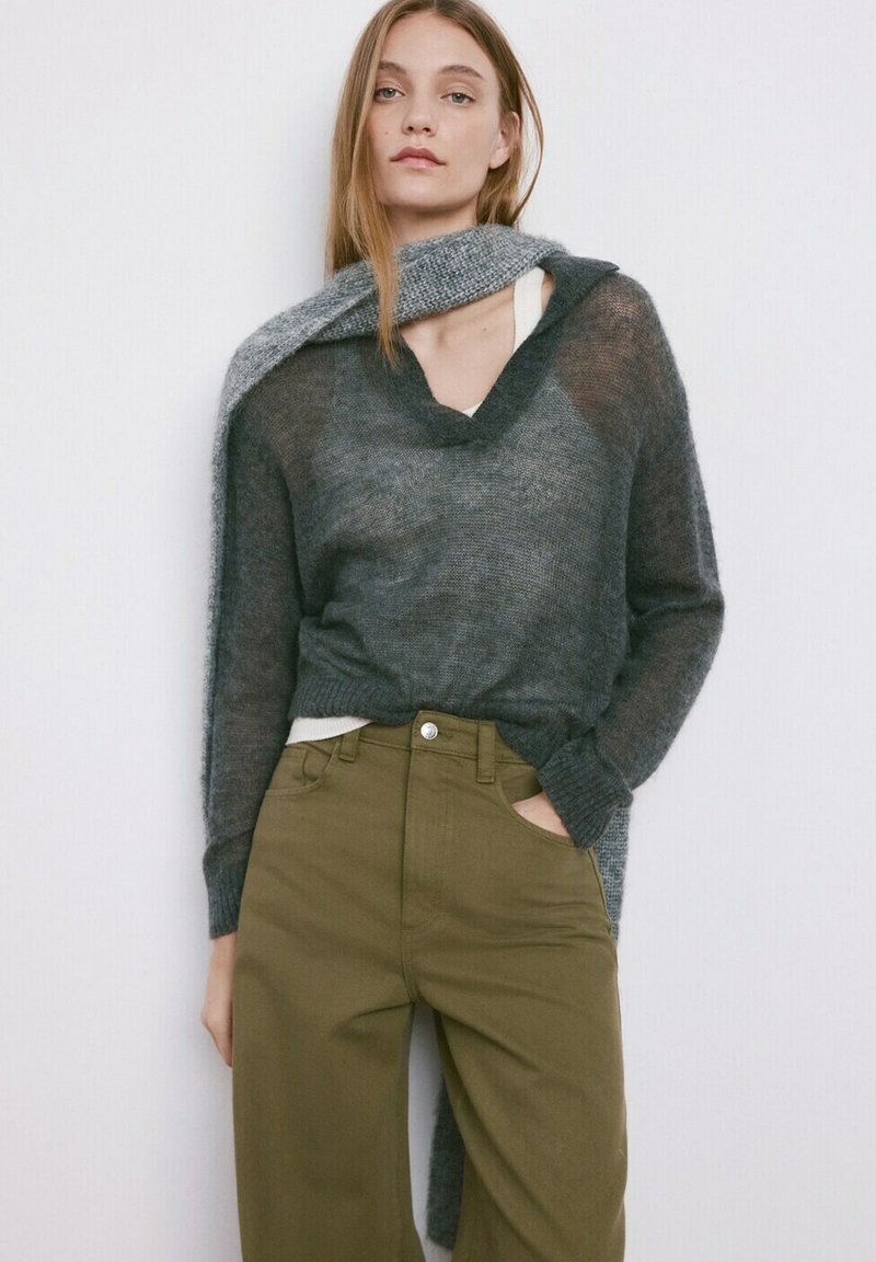 Femeie purtând un pulover din tricot transparent, de culoare gri închis, cu o eșarfă asortată drapată peste umeri, pantaloni kaki cu talie înaltă, stând în picioare lângă un perete alb.