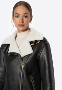 Czarna skórzana kurtka z puszystym białym kołnierzem, złotymi akcentami, zamkami błyskawicznymi oraz bocznymi kieszeniami. Gładka tekstura i dopasowany krój.