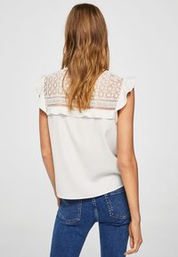 Blouse blanche avec des détails en dentelle dans le dos, manches à volants, et texture douce, associée à un jean bleu ajusté.