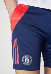 Námornícke športové šortky s červeným gradientom, s logom Manchester United na dolnej ľavej nohe a tromi červenými pruhmi na boku.