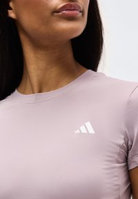 Κοντινό πλάνο ενός ατόμου που φοράει αθλητική μπλούζα ανοιχτό μωβ με λευκό λογότυπο Adidas στο στήθος, εμφανίζοντας τον λαιμό και μέρος του προσώπου.