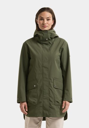 Frau trägt eine lange olivgrüne Kapuzen-Regenjacke mit Druckknopftaschen und beiger Hose, steht vor einem einfarbigen hellen Hintergrund.