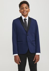 Giacca da abito blu navy con rever a scialle in satin nero, due tasche frontali e chiusura a un bottone. Abbinata a una camicia bianca e una cravatta nera.