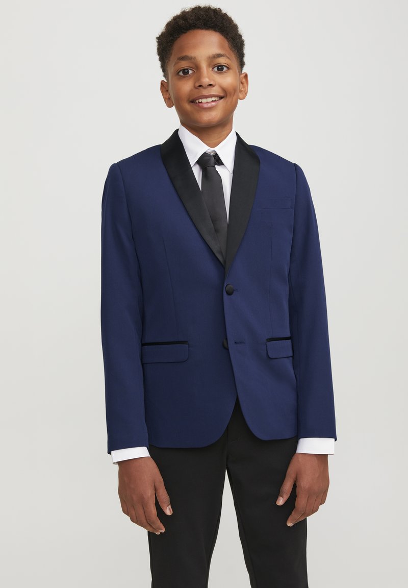 Giacca da abito blu navy con rever a scialle in satin nero, due tasche frontali e chiusura a un bottone. Abbinata a una camicia bianca e una cravatta nera.