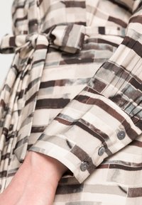 Blusa riscada em tecido suave com padrões em bege, cinza e marrom. O punho tem três botões redondos e um ligeiro franzido. Detalhe na cintura amarrada.
