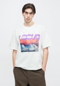 Fehér pamut póló, színes grafikával, amely felhőket és naplementét ábrázol. A felirat lilával "FLY ABOVE" (Repülj felett). Laza fazon, rövid ujjú.