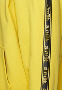 Yellow hoodie featuring a soft texture, kangaroo pocket, and a side stripe with "silverTab" branding in black and yellow.