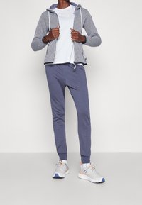 Sweatshirtjacka med ett geometriskt mönster i marinblått och vitt, tillsammans med grå sweatpants och ljusgrå sneakers med orange detaljer.