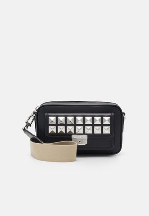 Sac bandoulière rectangulaire noir avec des clous pyramidaux argentés sur la poche avant et une large sangle beige en tissu.