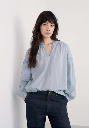 Vrouw met lang donker haar, gekleed in een lichtblauwe blouse met gathermouwen en donkere denim jeans, staand met handen in de zakken tegen een witte muur.