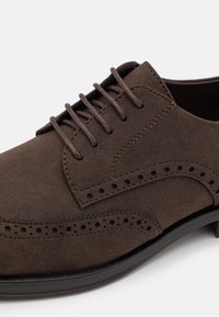 Brune semsket snøresko med brogue-detaljer, med en glatt tekstur og gummisåle, designet for formelle eller semi-formelle anledninger.