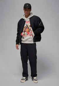 Zwarte bomberjack over een grijze hoodie met een grafische print, gecombineerd met zwarte cargo broeken en sneakers met grijs en creme accenten.