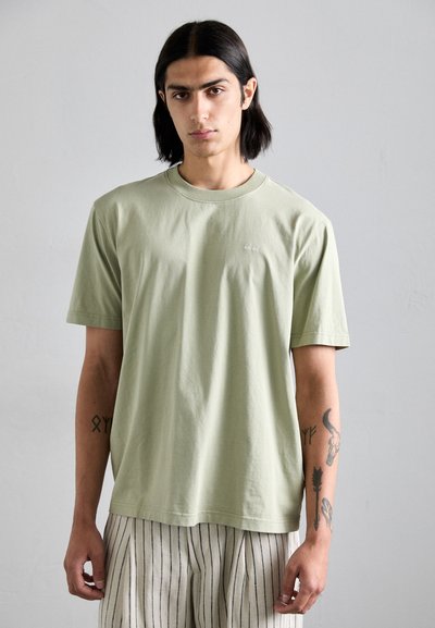 Ανοιχτό πράσινο βαμβακερό t-shirt κοντό μανίκι με λαιμόκοψη και μικρή λεπτομέρεια λογότυπου στο αριστερό στήθος. Το μοντέλο φοράει ριγέ παντελόνι.