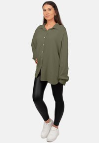 Olijfgroen, oversized button-up shirt van gestructureerde stof, lange mouwen en een ronde zoom, gecombineerd met zwarte leggings en witte sneakers.