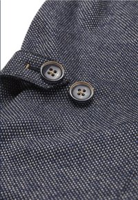 Marinblå textil med struktur som har två svarta, runda knappar vid kragen. Mönstret har en subtil vävning med nyanser av blått och grått.