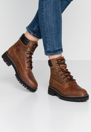Bottines montantes en cuir marron embossé avec col matelassé noir et semelles noires robustes, portées avec un jean bleu.