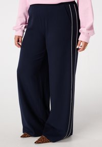 Marineblauwe wijde broek met verticale strepen, voorzien van zijzakken en een contrasterende witte bies. Zacht, gestructureerd materiaal. Leopardenprint hakken.