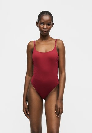 Femeie cu părul împletit, purtând un costum de baie întreg, fără mâneci, de culoare roșu închis, stând pe un fundal simplu gri deschis.