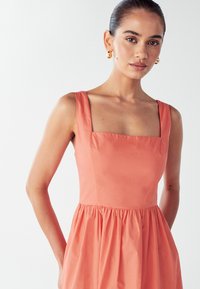 Koraljna haljina bez rukava s četvrtastim izrezom, prilagođenim korzetom i naboranom suknjom. Izrađena od glatkog materijala, s minimalnim šavovima i detaljima.