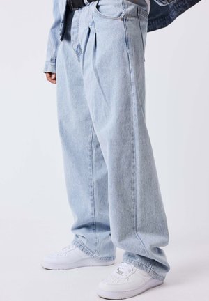 Personne portant un jean bleu clair à jambes larges avec des plis et des baskets blanches, debout devant un fond clair uni.