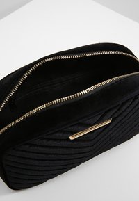 Černá prošívaná sametová kabelka s kovaným zipem a obdélníkovým zlatým kovovým detailem, otevřená, aby ukázala vnitřní kapsu na zip.