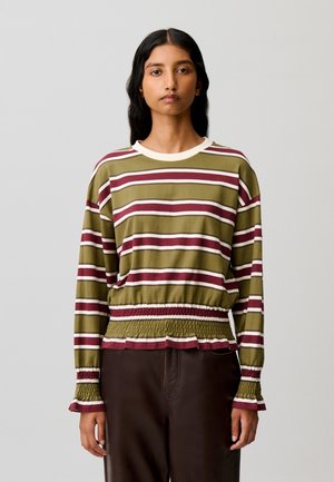 Donna con capelli lunghi e scuri che indossa una maglia a righe verde oliva, bordeaux e bianco con maniche lunghe e polsini e vita arricciati, in piedi davanti a uno sfondo semplice.