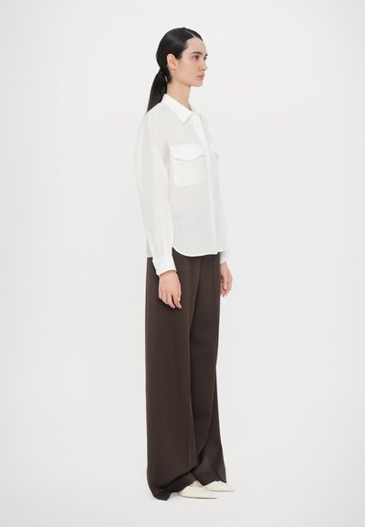 White long-sleeve shirt with two chest pockets, paired with high-waisted, wide-leg brown trousers. Smooth texture, minimal design.