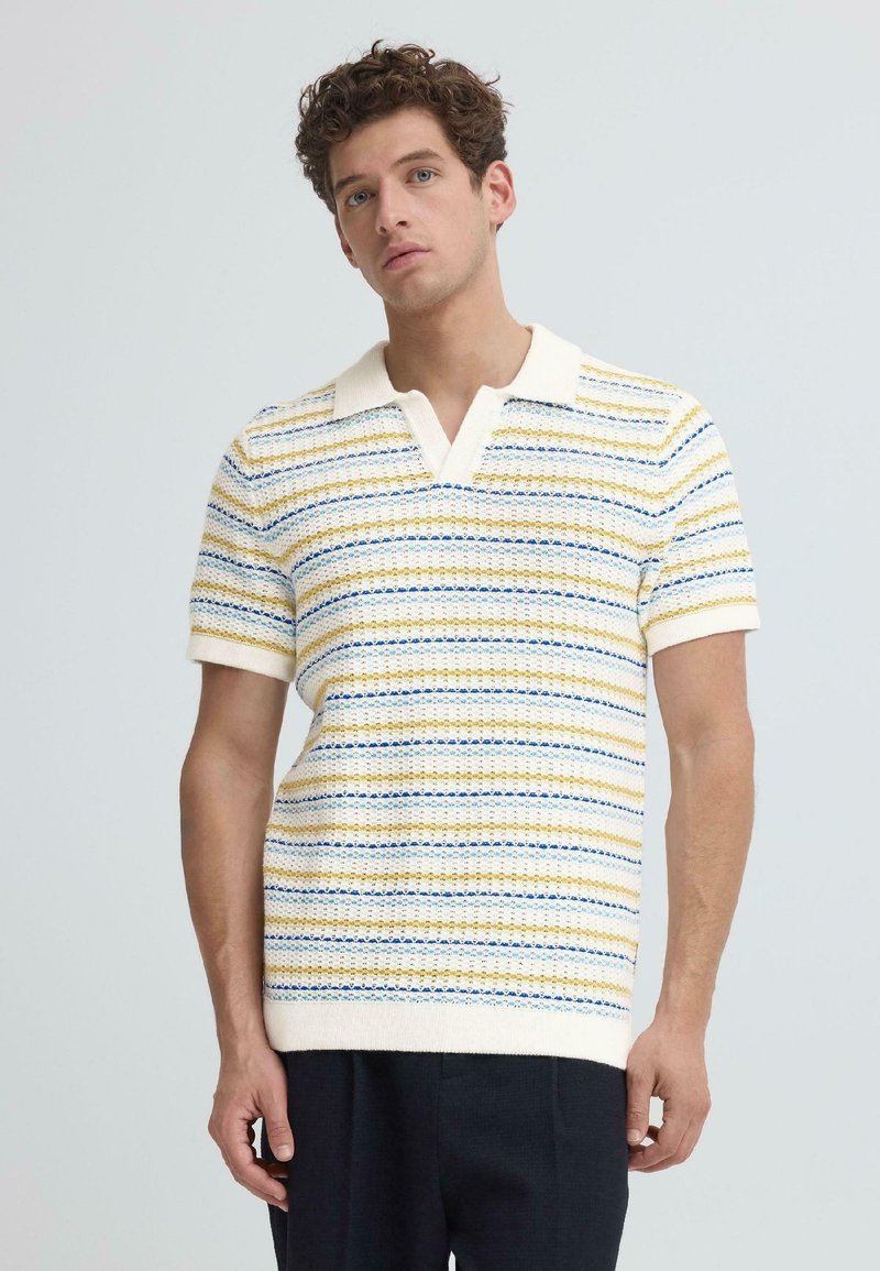Homme portant un polo en maille à manches courtes avec des rayures horizontales bleues, jaunes et blanches, associé à un pantalon foncé sur un fond clair uni.